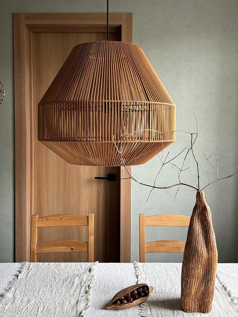 May include: A large, woven pendant light fixture hangs above a table. The light is made of tan-colored rope and has a geometric shape. A wooden vase with branches and a decorative wooden pod are on the table. Two wooden chairs are visible.