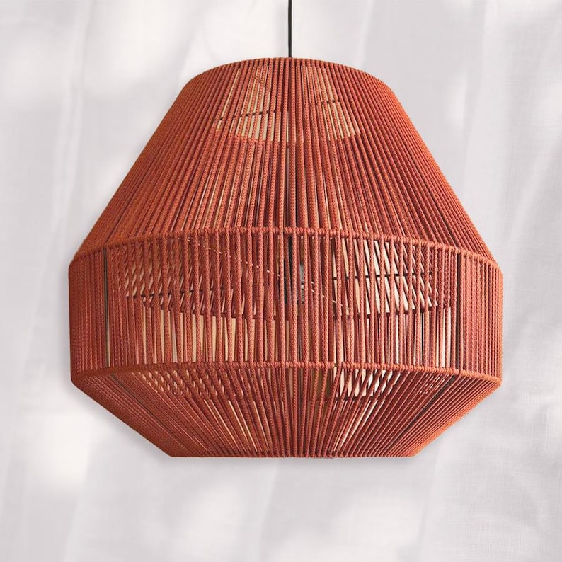 May include: A hanging pendant light fixture with a geometric design. The shade is made of rust-colored rope, creating a textured, open weave. The light fixture has a hexagonal shape and is suspended from a black cord.