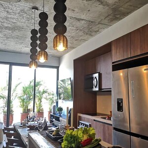 May include: Three hanging pendant lights with a stacked sphere design. The lights are black with a woven texture and a warm glow from the bulbs. The kitchen features wood cabinets, stainless steel appliances, and a concrete ceiling.
