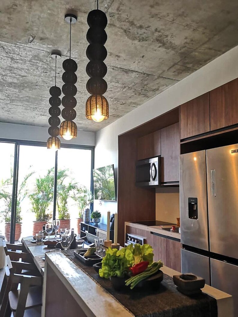 May include: Three hanging pendant lights with a stacked sphere design. The lights are black with a woven texture and a warm glow from the bulbs. The kitchen features wood cabinets, stainless steel appliances, and a concrete ceiling.