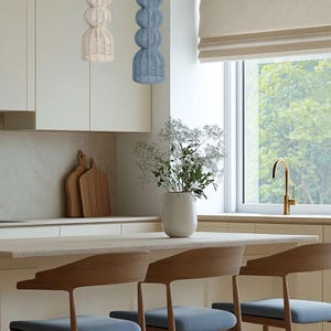 May include: A modern kitchen scene featuring two woven pendant lights, one cream and one blue, hanging above a countertop. Three wooden bar stools with blue seats are positioned in front of the counter. A vase of white flowers sits on the counter.