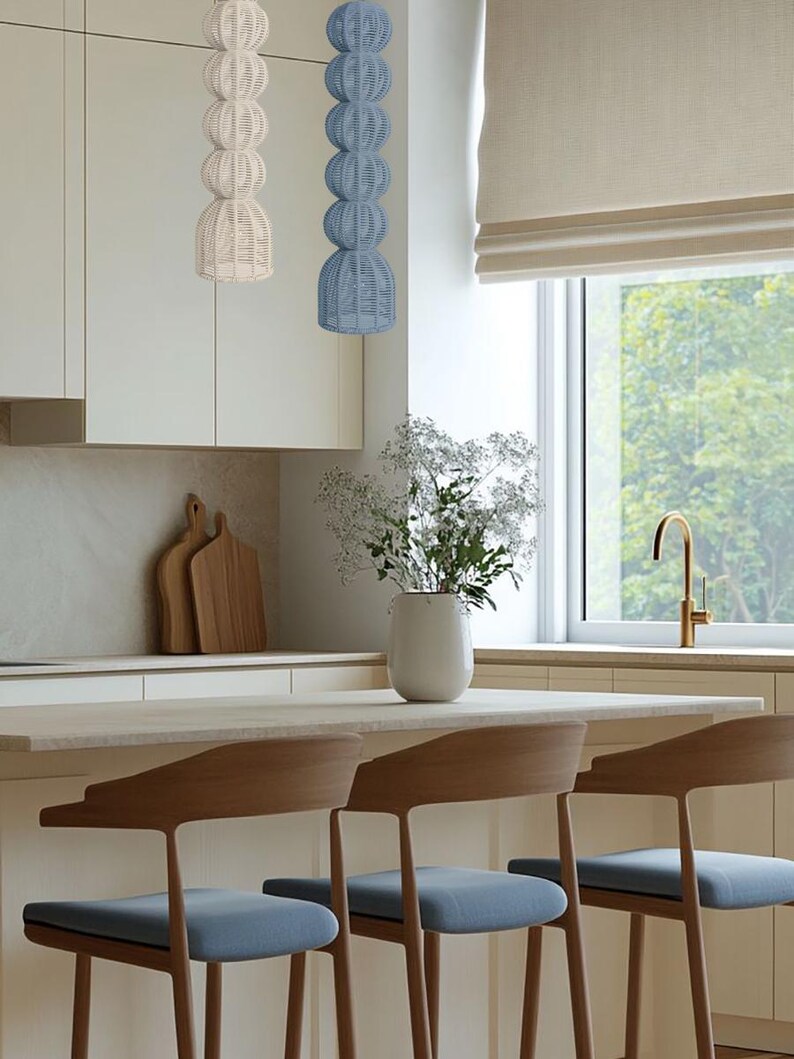 May include: A modern kitchen scene featuring two woven pendant lights, one cream and one blue, hanging above a countertop. Three wooden bar stools with blue seats are positioned in front of the counter. A vase of white flowers sits on the counter.