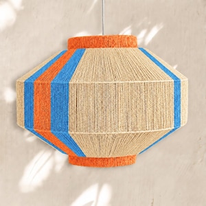 Può includere: Un paralume a sospensione di forma esagonale avvolto in spago beige naturale. Il design presenta strisce verticali di filo arancione brillante e blu, con fasce arancioni solide sui bordi superiore e inferiore.