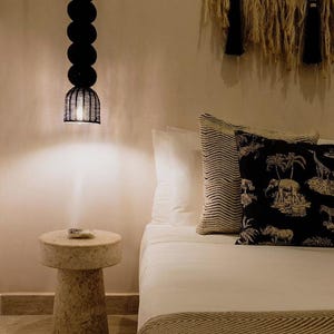 May include: A bedroom scene featuring a black pendant light with a series of round shades and a woven cage-like bottom. A small, round stone side table sits beside a bed with white linens and decorative pillows. A woven wall hanging adds texture.