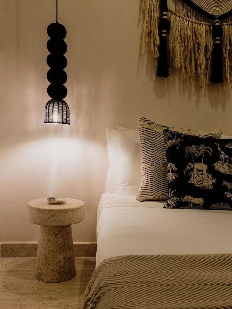 May include: A bedroom scene featuring a black pendant light with a series of round shades and a woven cage-like bottom. A small, round stone side table sits beside a bed with white linens and decorative pillows. A woven wall hanging adds texture.