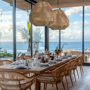 May include: A dining room set with a long wooden table and woven chairs. Two large, woven pendant lights hang above the table. The room has large windows with a view of the ocean.