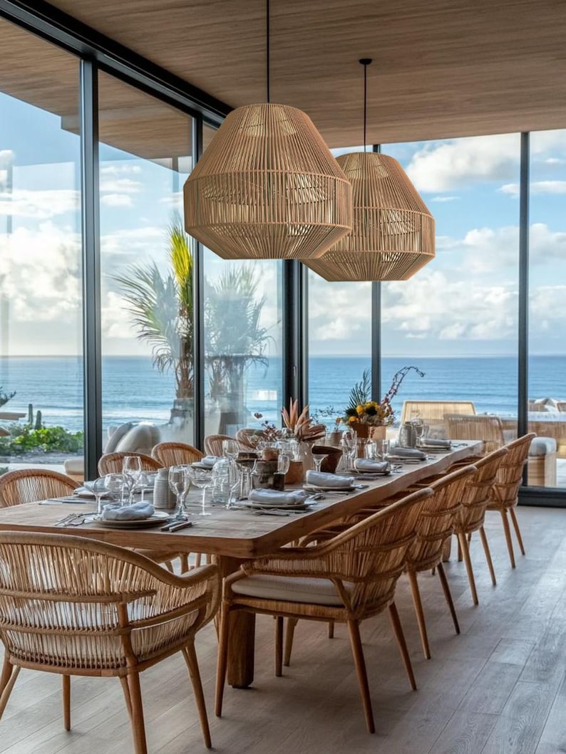 May include: A dining room set with a long wooden table and woven chairs. Two large, woven pendant lights hang above the table. The room has large windows with a view of the ocean.