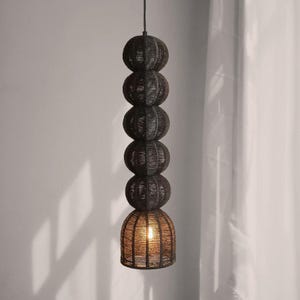 May include: A dark brown pendant light fixture with a stacked sphere design. The light has five woven spheres, with the bottom sphere shaped like a bell. The light is suspended from a black cord and emits a warm glow.