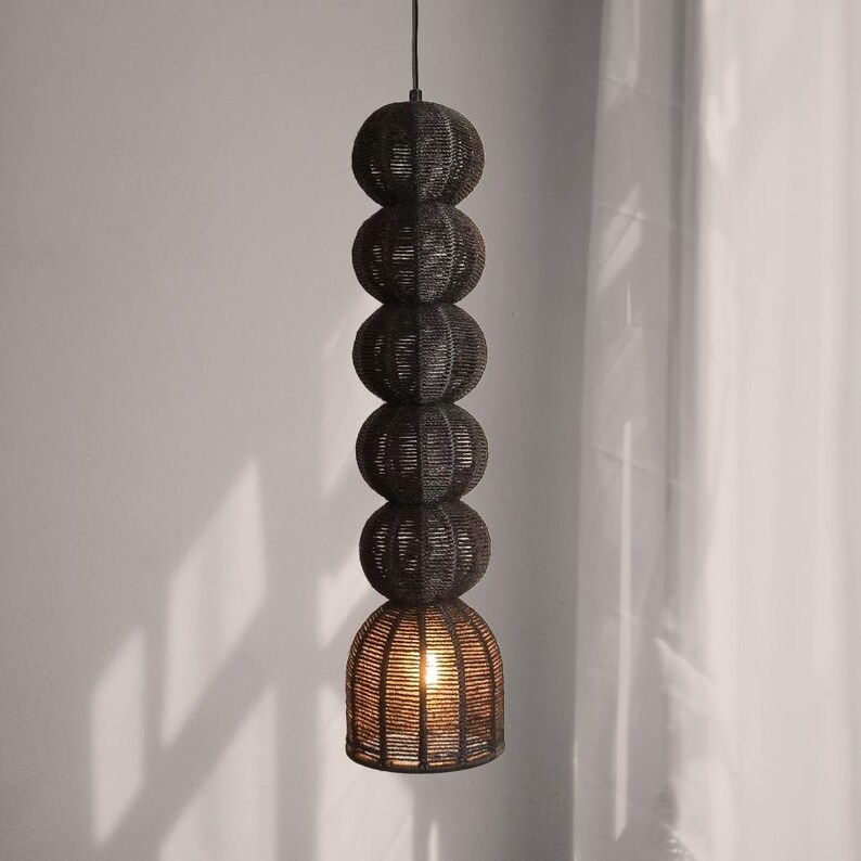 May include: A dark brown pendant light fixture with a stacked sphere design. The light has five woven spheres, with the bottom sphere shaped like a bell. The light is suspended from a black cord and emits a warm glow.