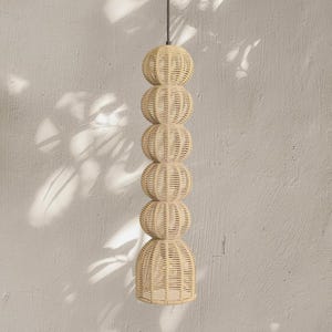 May include: A woven pendant light fixture with a stacked design. The light is made of natural-colored wicker and features a series of spherical shapes leading to a flared base. The fixture hangs from a black cord against a neutral wall.