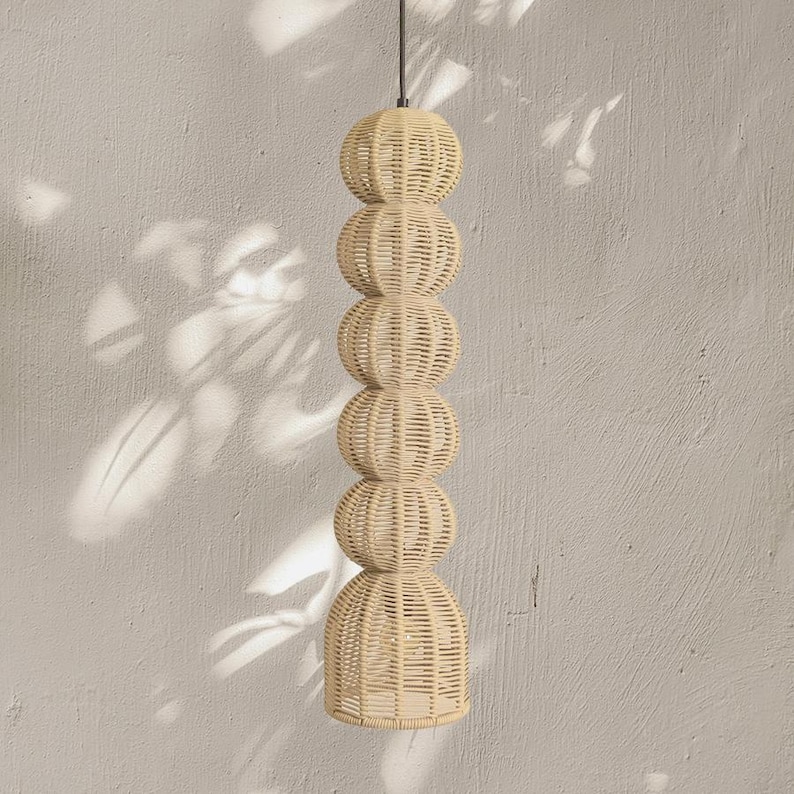 May include: A woven pendant light fixture with a stacked design. The light is made of natural-colored wicker and features a series of spherical shapes leading to a flared base. The fixture hangs from a black cord against a neutral wall.