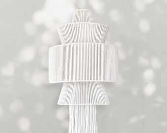Op de afbeelding: Een witte, gelaagde hanglamp. De lamp heeft een conische bovenkant, een breed cilindrisch middengedeelte en een uitlopende basis met hangende franjes. De armatuur is gemaakt van geweven materiaal, waardoor het een textuur krijgt. Het hangt aan een enkel snoer.