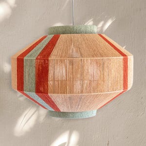 May include: A hanging pendant light fixture with a geometric shape. The light is made of woven natural-colored material with vertical stripes of red and green. The top and bottom are light green.