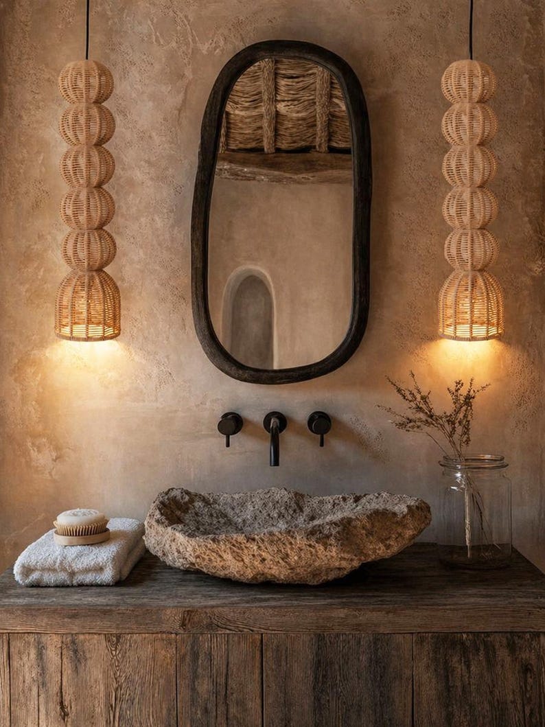 May include: A rustic bathroom scene featuring a stone sink, a dark wooden vanity, and a large oval mirror. Two woven pendant lights hang on either side of the mirror. The color palette is neutral with warm tones.
