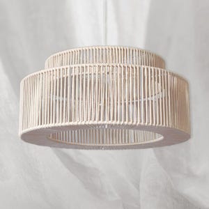 Może przedstawiać: Kremowa lampa wisząca z plecionej liny. Lampa ma warstwową konstrukcję z dużym, otwartym dolnym pierścieniem i mniejszym górnym pierścieniem. Pionowe liny tworzą teksturowany, cylindryczny klosz.