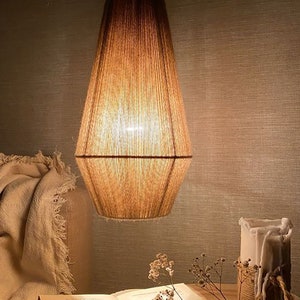 Può includere: Una lampada a sospensione con un paralume marrone, strutturato e allungato. La luce è sospesa da un cavo nero. Il paralume è illuminato dall'interno. Sotto, un libro aperto, una candela e fiori secchi sono visibili. La lampada è un elemento decorativo.