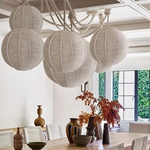 May include: A collection of white, spherical pendant lights with a woven texture hangs above a dining table. The lights are suspended from a rope-like fixture. The dining table is set with various decorative vases and bowls.