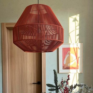 May include: A hanging, red-orange pendant light fixture with a woven rope design. The light has a geometric shape and is suspended from a black cord. The fixture hangs above a wooden table with a floral arrangement.