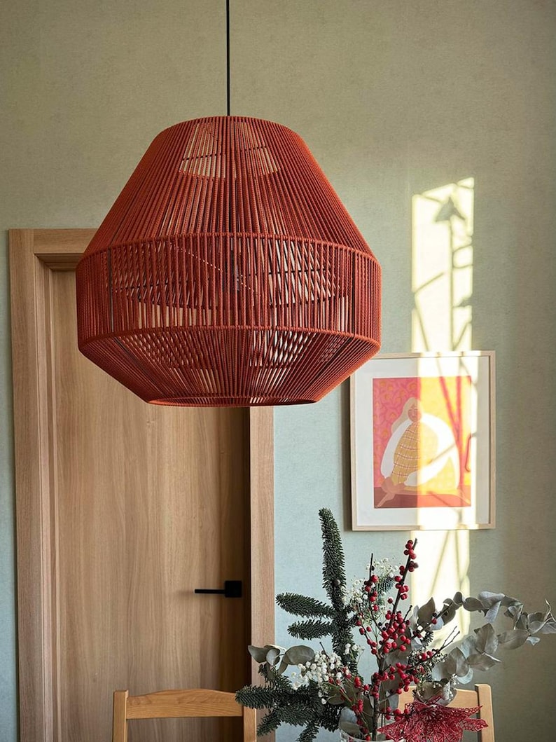 May include: A hanging, red-orange pendant light fixture with a woven rope design. The light has a geometric shape and is suspended from a black cord. The fixture hangs above a wooden table with a floral arrangement.