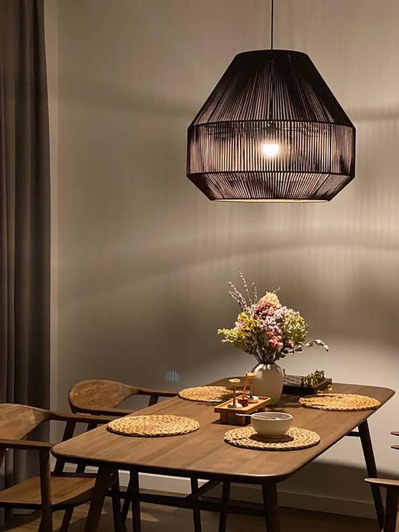 May include: A dark brown, geometric pendant light hangs above a wooden dining table. The table is set with woven placemats, a vase of flowers, and a small bowl. Wooden chairs surround the table, creating a warm and inviting dining space.