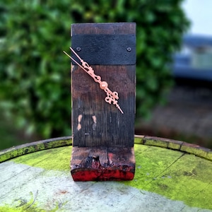 Könnte beinhalten: Eine rustikale Holzuhr mit einem schwarzen Metallzifferblatt und kupfernen Zeigern. Die Uhr ist auf einem Holzsockel montiert und hat eine abgenutzte Oberfläche.