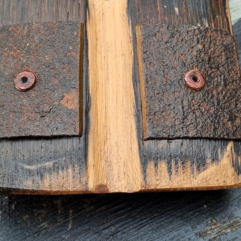 May include: Close-up of a wooden object with a central, light-colored section flanked by darker, textured panels. Copper-colored rivets secure the panels. The wood grain is visible, and the overall appearance is rustic and handcrafted.