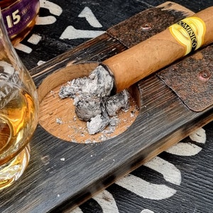 May include: A lit cigar with the label "Dominico" rests on a wooden ashtray. The ashtray is part of a larger wooden serving tray. A glass of amber liquid is in the background.