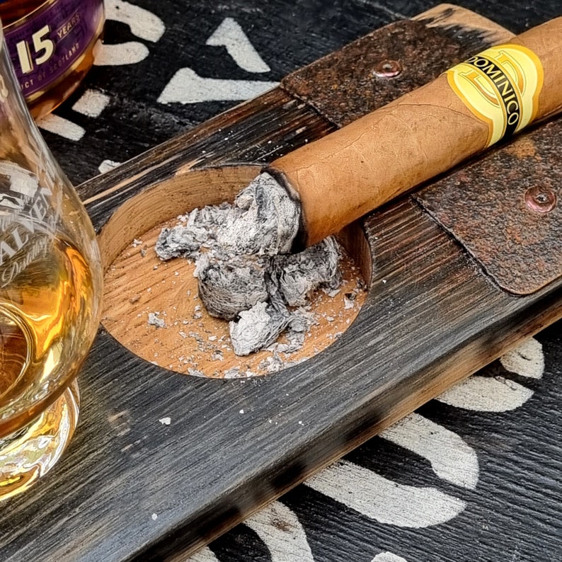 May include: A lit cigar with the label "Dominico" rests on a wooden ashtray. The ashtray is part of a larger wooden serving tray. A glass of amber liquid is in the background.