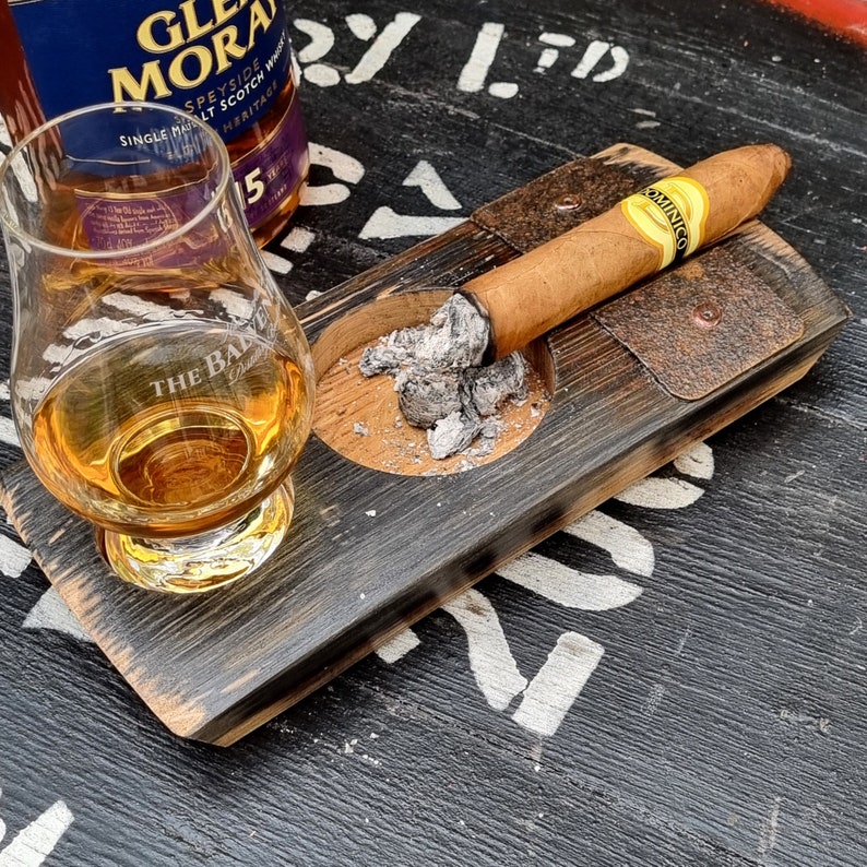 May include: A glass of amber liquid and a bottle of Glen Moray Speyside Single Malt Scotch Whisky sit next to a lit cigar resting on a wooden ashtray. The ashtray is made from a repurposed barrel stave.