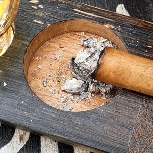 May include: A wooden ashtray with a circular indentation holding a lit cigar and ash. The ashtray is made of dark, weathered wood with a lighter wood interior. The cigar is a light brown color.