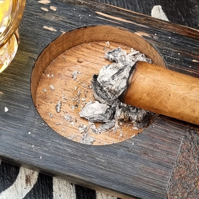 May include: A wooden ashtray with a circular indentation holding a lit cigar and ash. The ashtray is made of dark, weathered wood with a lighter wood interior. The cigar is a light brown color.