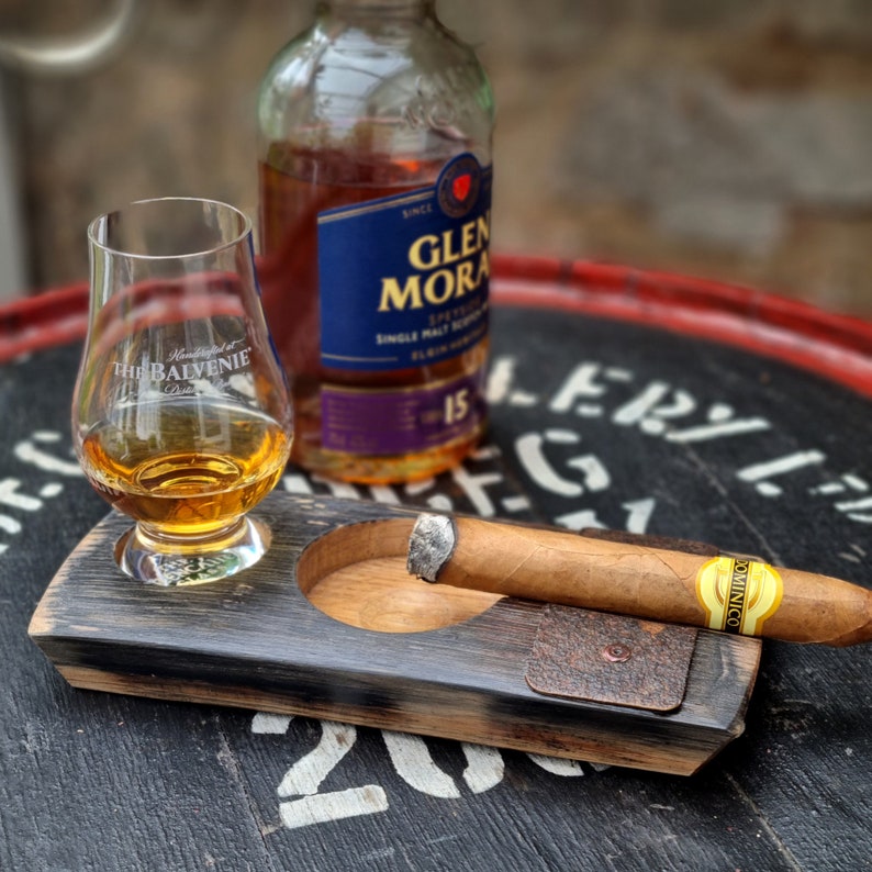May include: A whiskey glass with amber liquid, a bottle of Glen Moray Speyside Single Malt Scotch Whisky, and a lit cigar rest on a wooden ashtray. The ashtray has a small leather patch and a circular indentation for the glass.