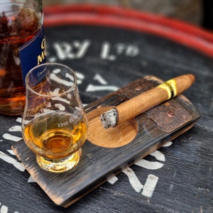 May include: A glass of amber-colored liquor and a lit cigar rest on a wooden tray. A bottle of liquor is partially visible in the background. The tray has a built-in ashtray and is made from a repurposed barrel.