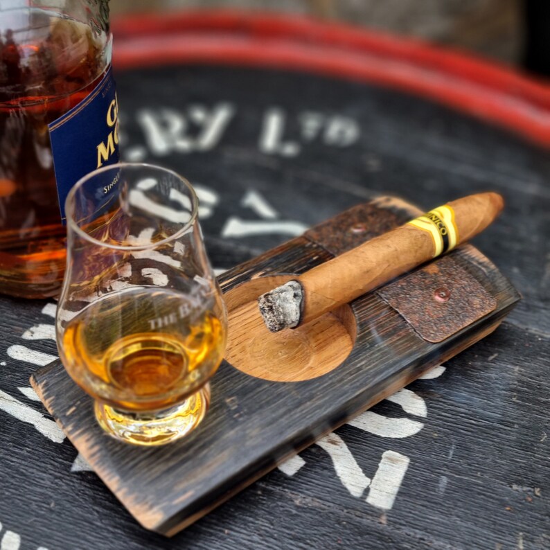 May include: A glass of amber-colored liquor and a lit cigar rest on a wooden tray. A bottle of liquor is partially visible in the background. The tray has a built-in ashtray and is made from a repurposed barrel.