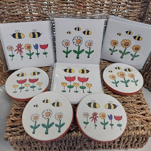 May include: Set of six coasters and six greeting cards featuring a whimsical bee and flower design. The coasters are round and have a white background with a red, yellow, and blue flower design. The greeting cards are white with a similar design.
