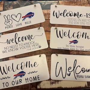 May include: A collection of white rectangular wooden signs with black text and various designs. Some signs feature the word "Welcome," while others display phrases like "Peace Love Bills" and "Welcome to our home."