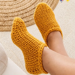 Op de afbeelding: Mosterdgele gebreide pantoffels. De pantoffels hebben een dikke brei textuur en een geribbelde manchet. De afbeelding toont de pantoffels aan de voeten van een persoon, rustend op een wit oppervlak met een geweven mat op de achtergrond.