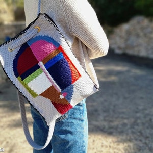 May include: A gray crochet shoulder bag with a colorful geometric design. The bag features a patchwork of pink, blue, red, green, and brown shapes. It has a black trim, gold eyelets, and a gray strap.