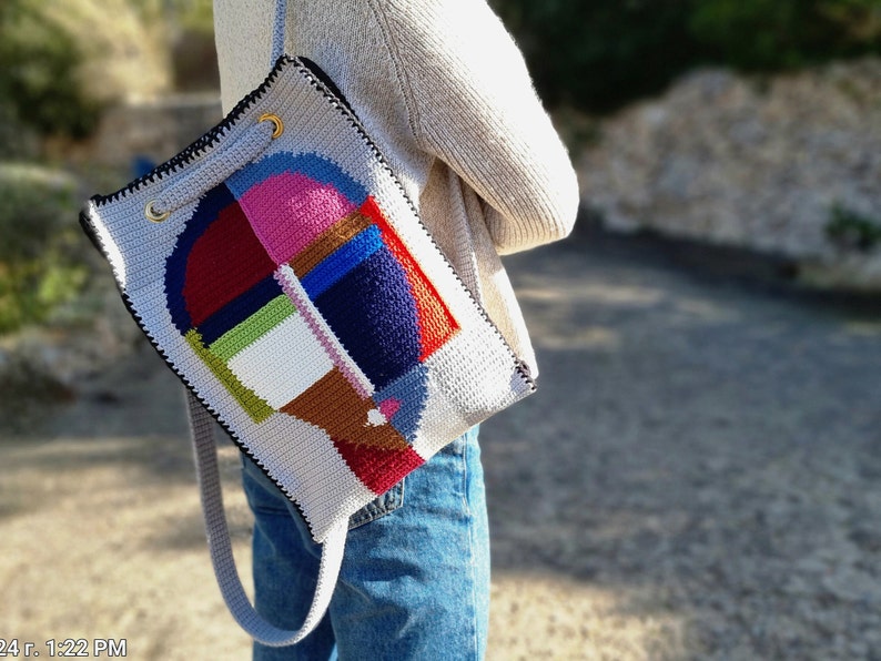 May include: A gray crochet shoulder bag with a colorful geometric design. The bag features a patchwork of pink, blue, red, green, and brown shapes. It has a black trim, gold eyelets, and a gray strap.