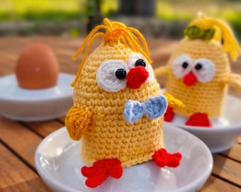 May include: Two yellow crocheted chick egg cozies featuring white eye patches, black pupils, red beaks, and red feet. One chick has a light blue bow tie, while the other has a green bow on its head. Both are placed on white ceramic saucers.