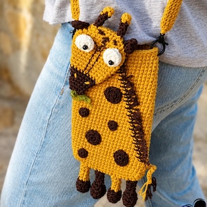 May include: A handmade, yellow crochet crossbody bag shaped like a giraffe. The bag features brown spots, a giraffe face with white eyes, and a long strap. The bag is worn over the shoulder.