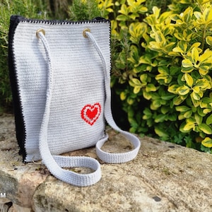 May include: A handmade crochet tote bag in light gray with a black border and straps. A red heart is embroidered on the front. The bag has gold-colored eyelets for the straps.
