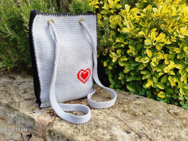 May include: A handmade crochet tote bag in light gray with a black border and straps. A red heart is embroidered on the front. The bag has gold-colored eyelets for the straps.
