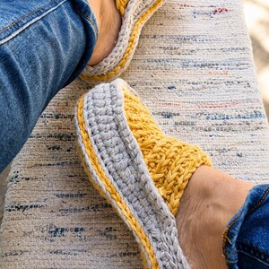 May include: A pair of handmade crochet slippers in yellow and gray. The slippers have a thick, textured design with a yellow top and gray sides, accented with a yellow border. The person is wearing blue jeans.
