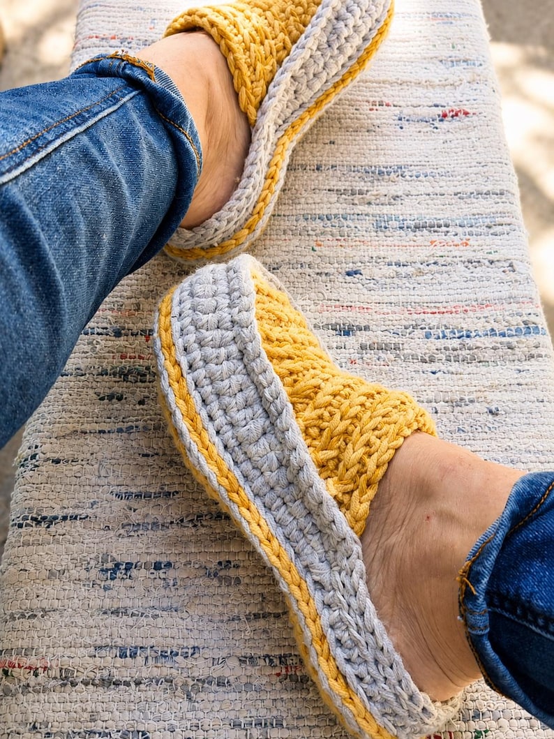 May include: A pair of handmade crochet slippers in yellow and gray. The slippers have a thick, textured design with a yellow top and gray sides, accented with a yellow border. The person is wearing blue jeans.