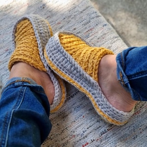May include: A pair of handmade crochet slippers. The slippers are yellow with a gray border and a yellow trim. The person wearing the slippers is wearing blue jeans.