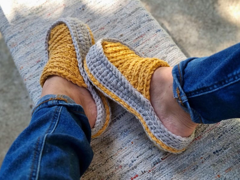 May include: A pair of handmade crochet slippers. The slippers are yellow with a gray border and a yellow trim. The person wearing the slippers is wearing blue jeans.