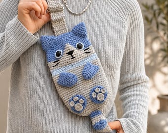 May include: A crochet crossbody bag shaped like a cat with a blue head, grey body, and blue paws. The cat features black embroidered whiskers, a small black nose, and round black eyes. A matching grey crochet strap is attached to the top.