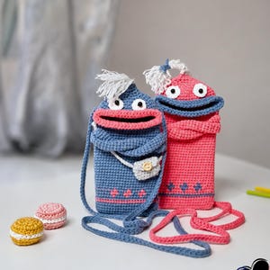 Könnte beinhalten: Zwei handgefertigte gehäkelte Handytaschen in Blau und Pink, jeweils mit einem skurrilen Monstergesicht-Design. Die blaue Tasche hat eine kleine weiße Geldbörse. Die Taschen haben lange Träger und sind mit weißen Haaren und einem rosa Mund verziert.