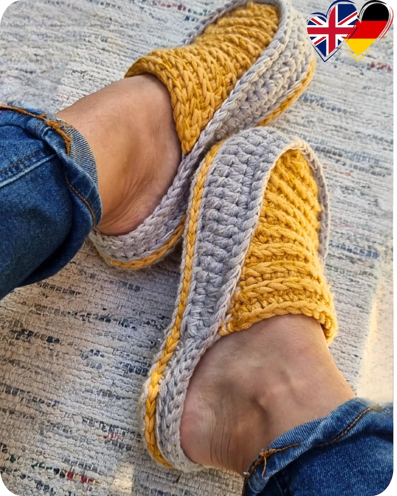 May include: A pair of handmade crochet slippers in yellow and gray. The slippers have a thick, textured appearance with a yellow top and a gray border. The person wearing the slippers is also wearing blue jeans.